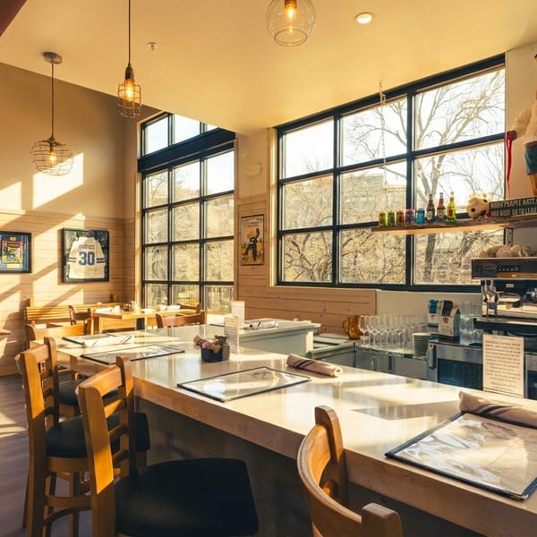 Bright, modern restaurant interior with a long bar, wooden tables, large windows, and a chef statue.