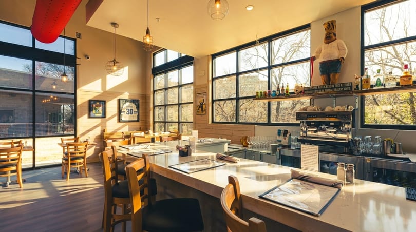 Bright, modern restaurant interior with a long bar, wooden tables, large windows, and a chef statue.