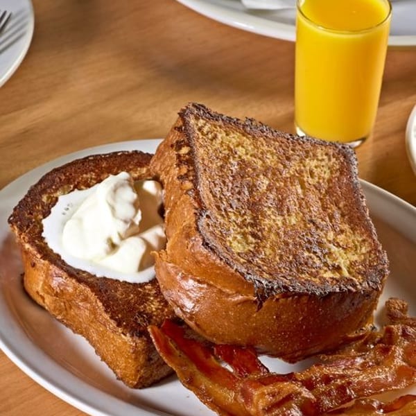 Thick-cut French toast with whipped cream and bacon, alongside orange juice and an omelet.