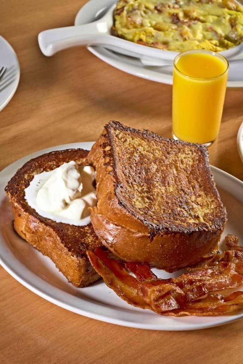 Thick-cut French toast with whipped cream and bacon, alongside orange juice and an omelet.