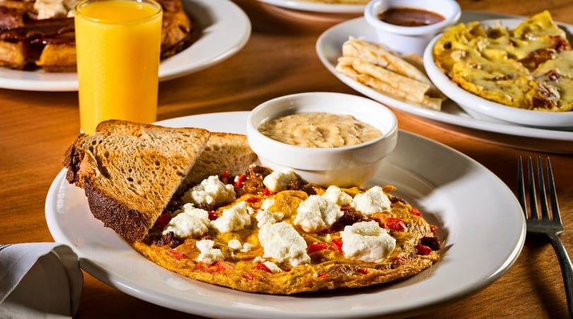 Breakfast spread with an omelet, toast, gravy, orange juice, and another omelet in the background.