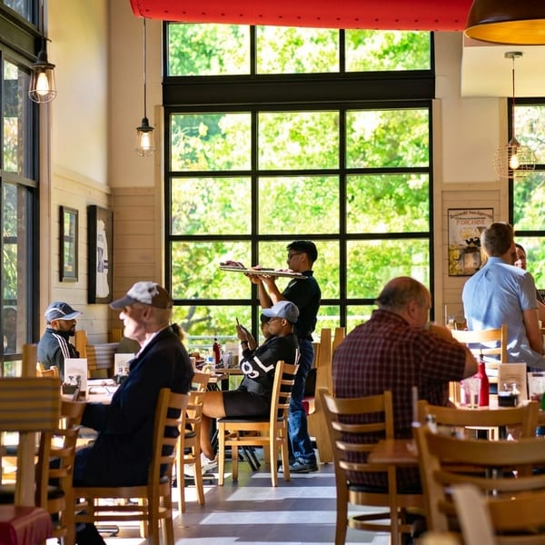 A bright restaurant dining room with large windows, customers seated at tables, and a server carrying food.