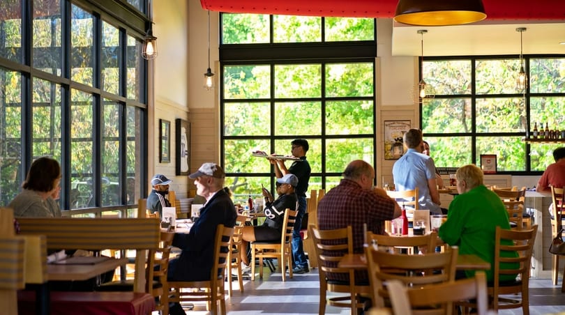 A bright restaurant dining room with large windows, customers seated at tables, and a server carrying food.