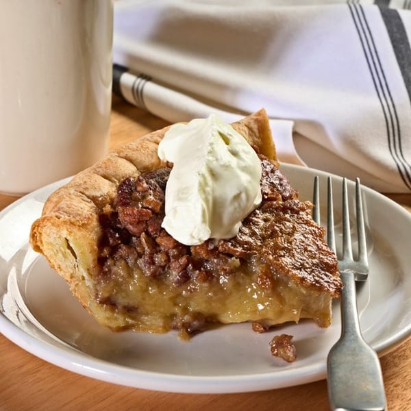 Slice of pecan pie with whipped cream on a white plate, fork, and coffee mug on a wooden table.