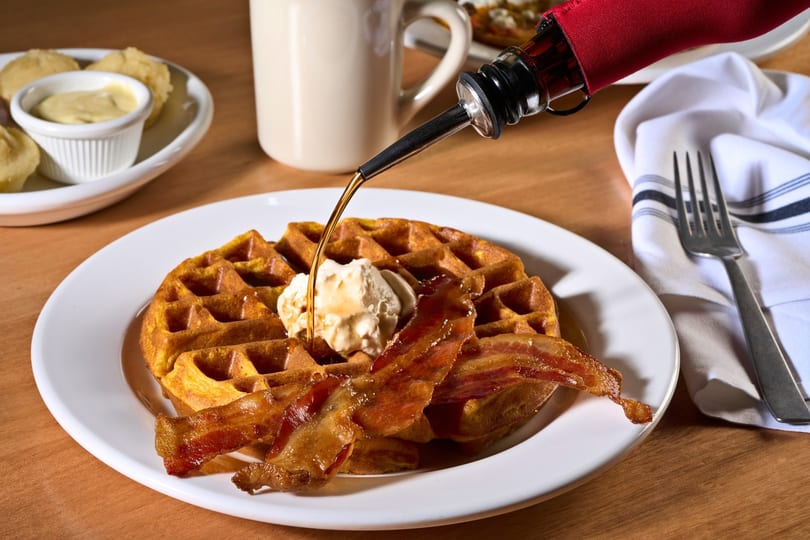 Maple syrup pouring onto a waffle with bacon and a scoop of butter on a white plate.