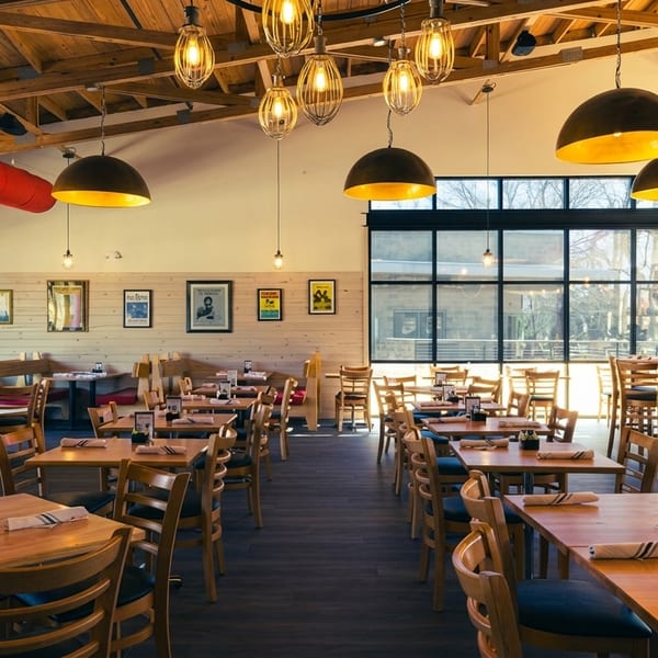 Spacious restaurant dining room with wooden tables, chairs, booths, and large windows, under warm pendant lights.