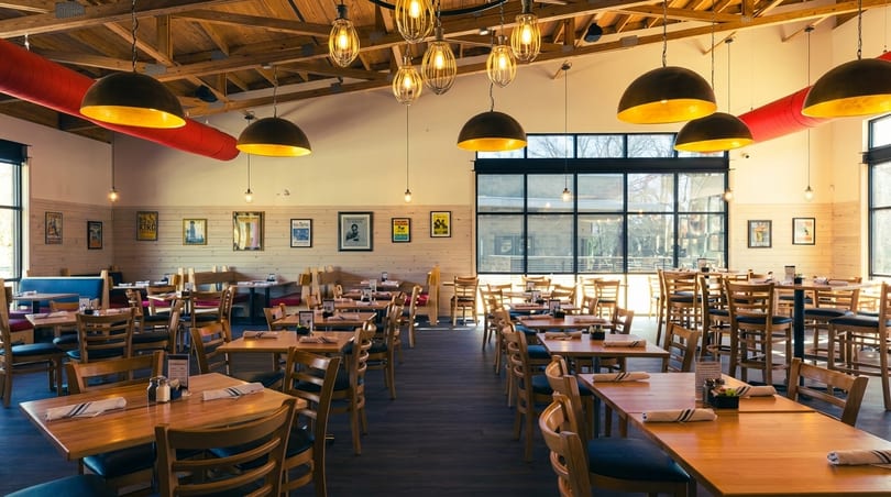 Spacious restaurant dining room with wooden tables, chairs, booths, and large windows, under warm pendant lights.
