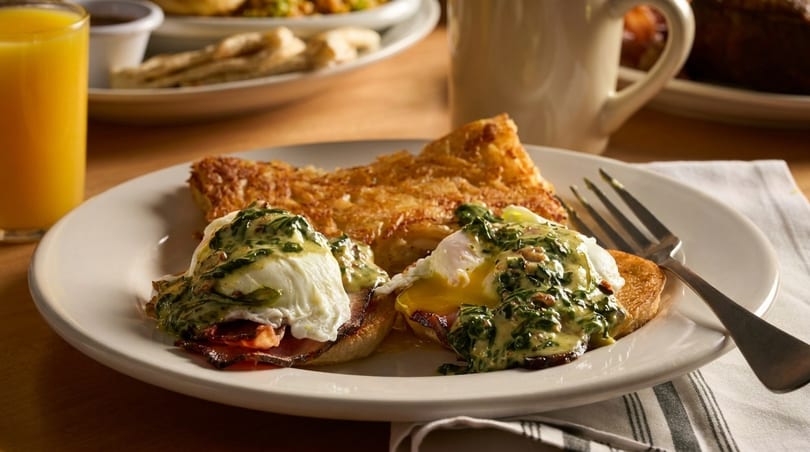 Eggs Benedict with spinach and hash browns, orange juice, and coffee on a wooden table.