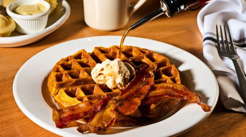 Syrup pouring onto a waffle with a scoop of butter and crispy bacon on a white plate.