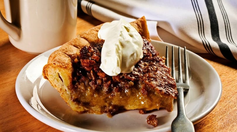 A slice of pecan pie with a dollop of whipped cream on a white plate with a fork.